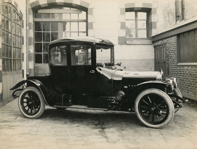Exposition Panhard-Levassor dans la fabrique de voitures d'Albert Lemaître, une voiture 12 HP coupé 2 places conduite intérieure à moteur 4 cylindres (1912), avenue Wilson, à proximité de l'atelier de menuiserie et du magasin de voitures, photographie noir et blanc, (15.6cm x 12cm), collection particulière établissements Lemaître, acquisition 2022, AMA.